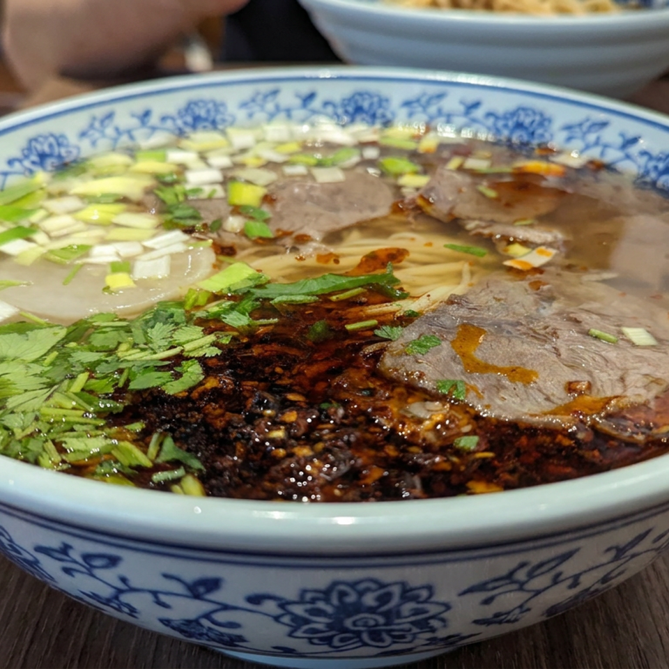 Large bowl of Chinese beef noodle soup topped with sliced beef, cilantro, scallions, and chili oil on a wooden table.