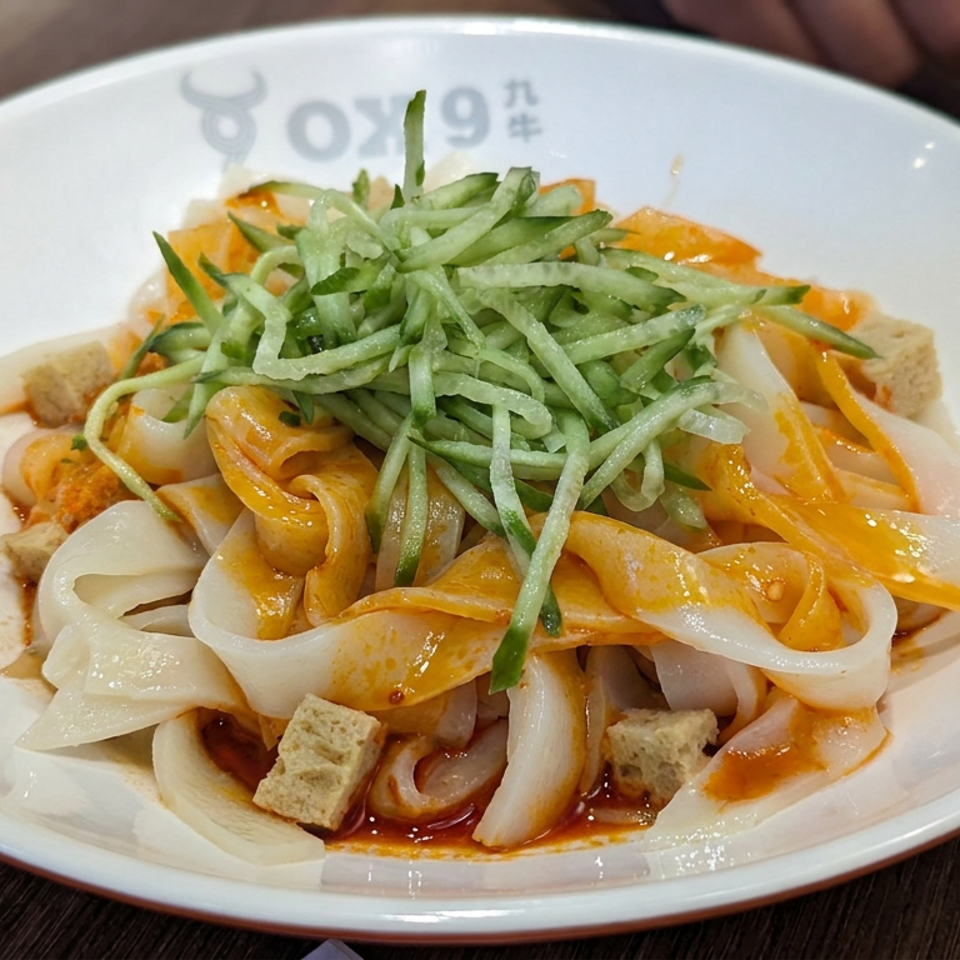 Wide rice noodles with tofu, vegetables, and shredded cucumber drizzled in orange sauce on a white plate.