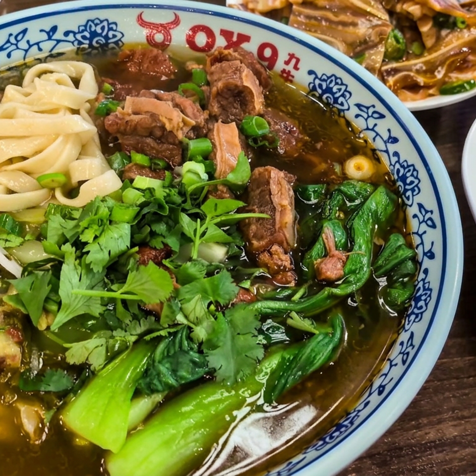 Bowl of beef noodle soup with wide noodles, leafy greens, cilantro, and scallions, alongside cucumber and vegetable dishes.